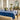 A set dining table with a blue tablecloth featuring a hydrangea pattern, surrounded by window light and decorative room elements.