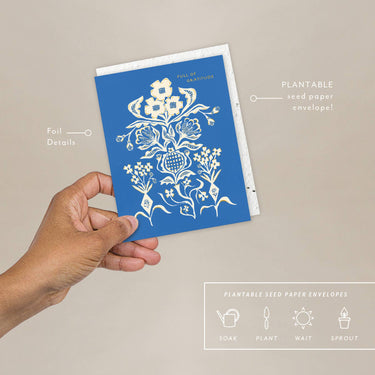 Hand holding a blue plantable seed paper envelope with white floral design on a beige background