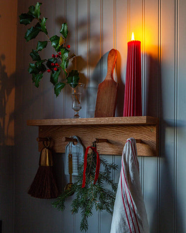 This image shows a shelf with hooks upon which sits a Greentree Petite Pillar candle in Red, lit. It sits amongst other winter items.