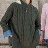 A woman wearing an oversized dark olive green cable-knit sweater over a light blue denim collared shirt, standing outdoors against a stone wall.