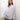 Brunette model is leaning up against a white wall. She is wearing a white button up top, light denim pants and is carrying an off-white cable-knit sweater.