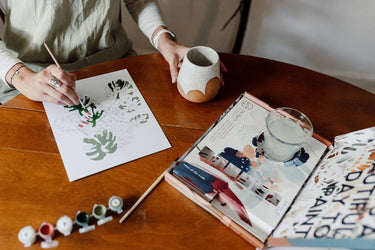 Person painting on paper with a cup of tea at a wooden table